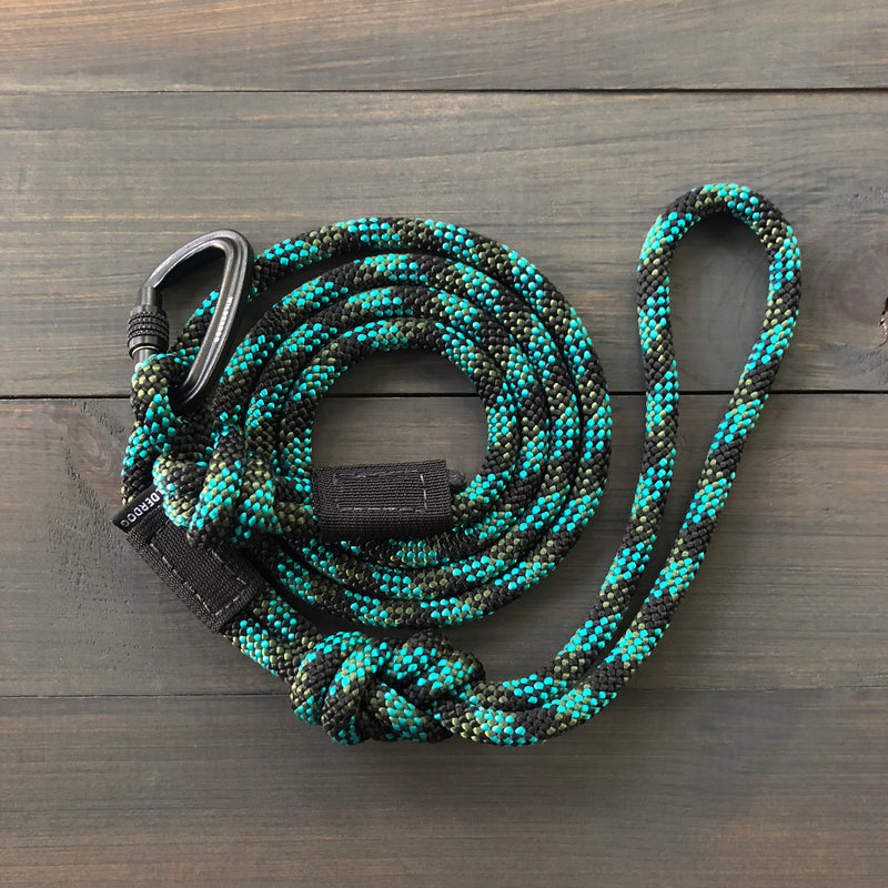 A meticulously coiled Granite Small Carabiner Leash, featuring a blue and black pattern with a loop at one end secured by a carabiner lock, is laid out on a wooden surface.
